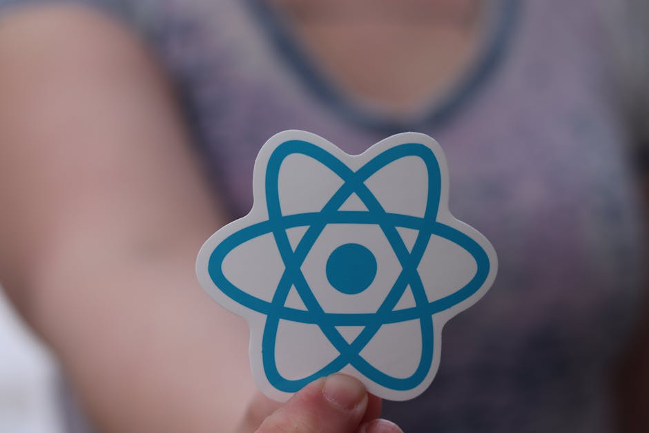 Close-up of a person's hand holding a sticker featuring a blue atomic model symbol in the foreground, with the person's blurred upper body and grey top visible behind. The background is out of focus, 