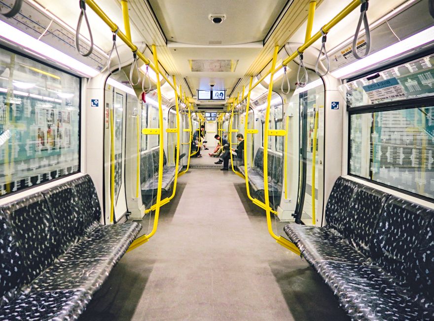 Interior view of a well-maintained train carriage with a clean, light-colored flooring and seats. The seats are upholstered in dark fabric with a patterned design, arranged in pairs along both sides o