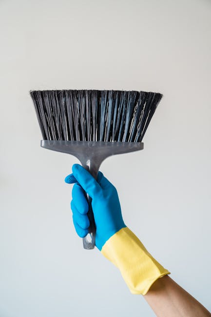 A person's hand wearing a blue glove and a yellow sleeve is holding a black dustpan and brush with stiff bristles against a plain white background, exemplifying surface cleaning tools used in domestic